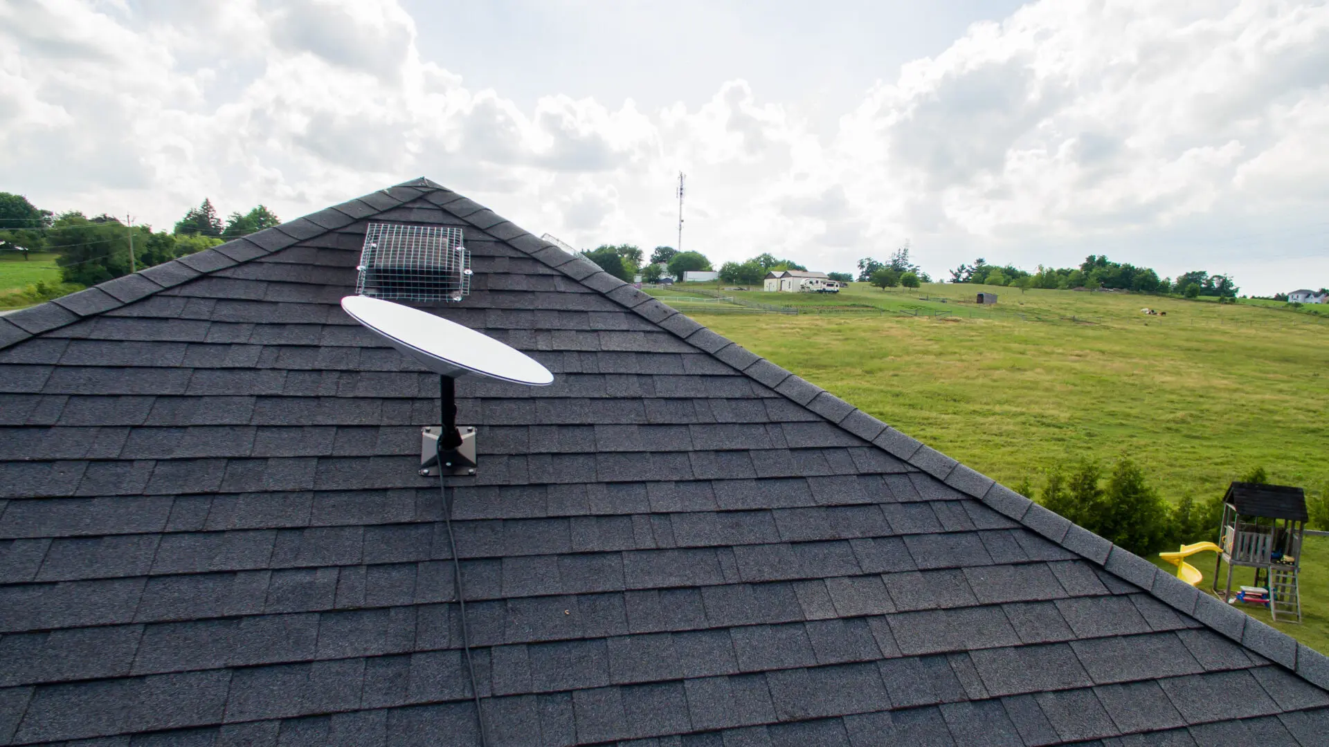 Satellite dish mounted on a shingled rooftop with a grassy hill in the background.
