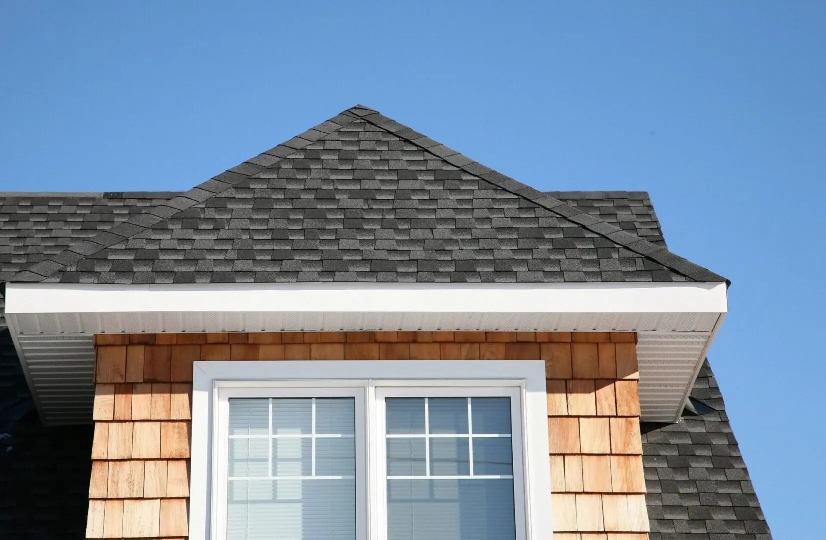 A house roof with wooden shingles and a window beneath a clear blue sky.